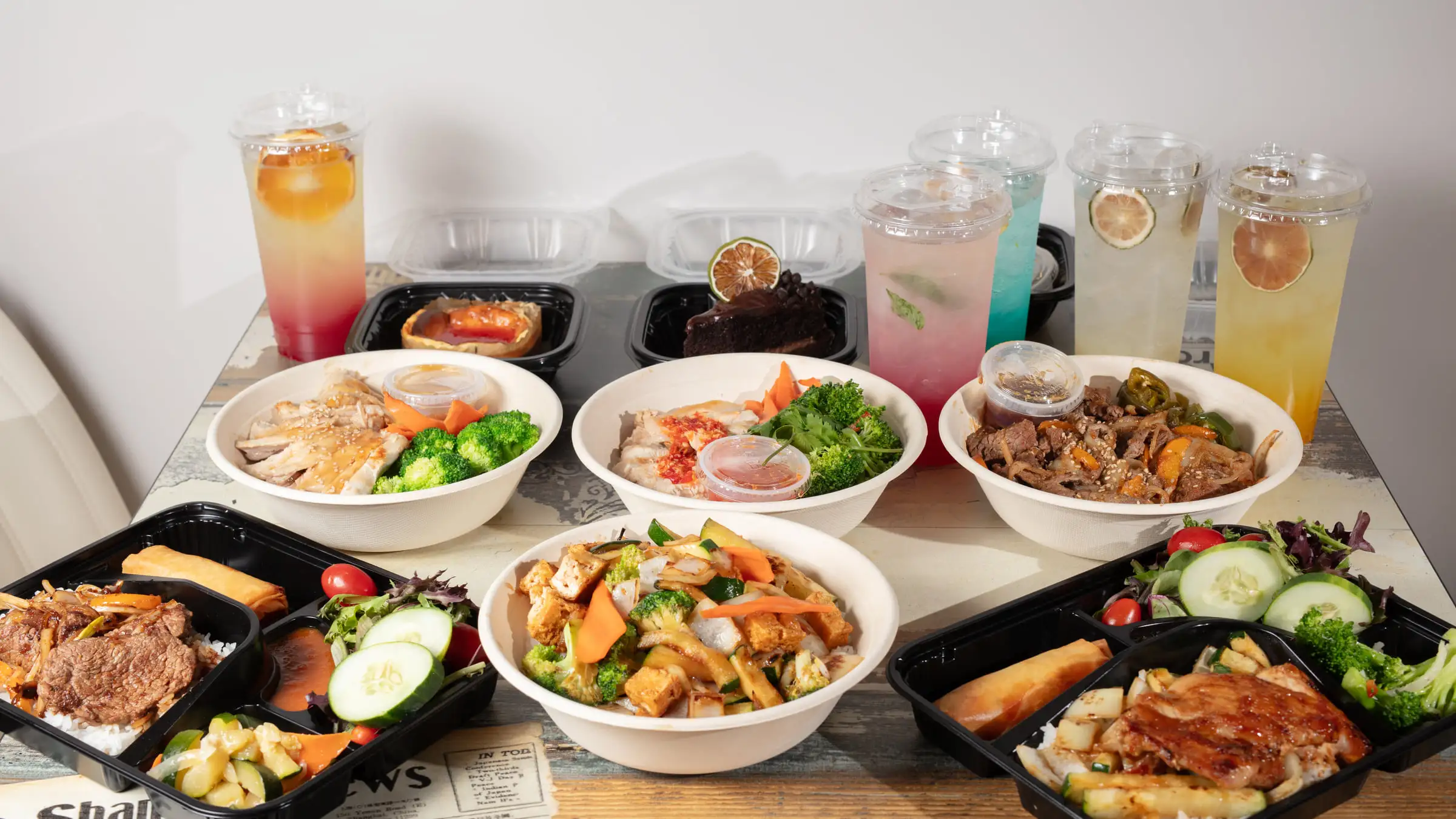 A spread of hibachi bowls, black bento trays and colored house drinks laid out together on a pale wooden table